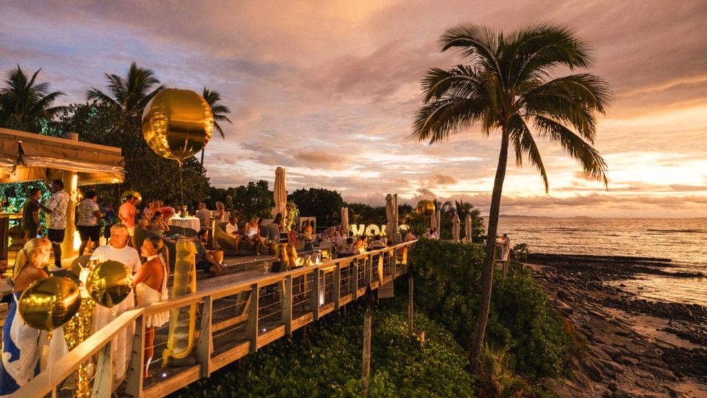 People dining on an outdoor wooden deck overlooking the ocean at sunset.