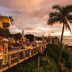 People dining on an outdoor wooden deck overlooking the ocean at sunset.