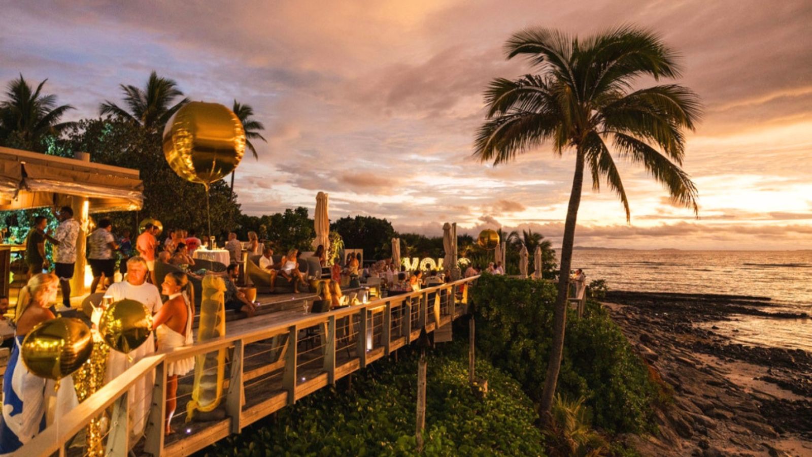 People dining on an outdoor wooden deck overlooking the ocean at sunset.
