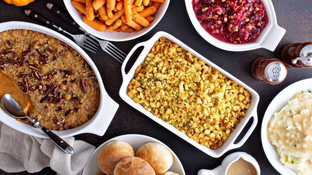 Overhead view of Thanksgiving side dishes including stuffing, sweet potato casserole, carrots, cranberry sauce, rolls, and mashed potatoes with gravy.