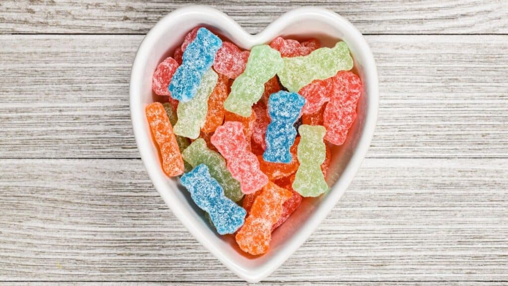 A heart-shaped bowl filled with colorful, sugar-coated gummy candies, reminiscent of the oldest Halloween candies, set on a wooden surface.