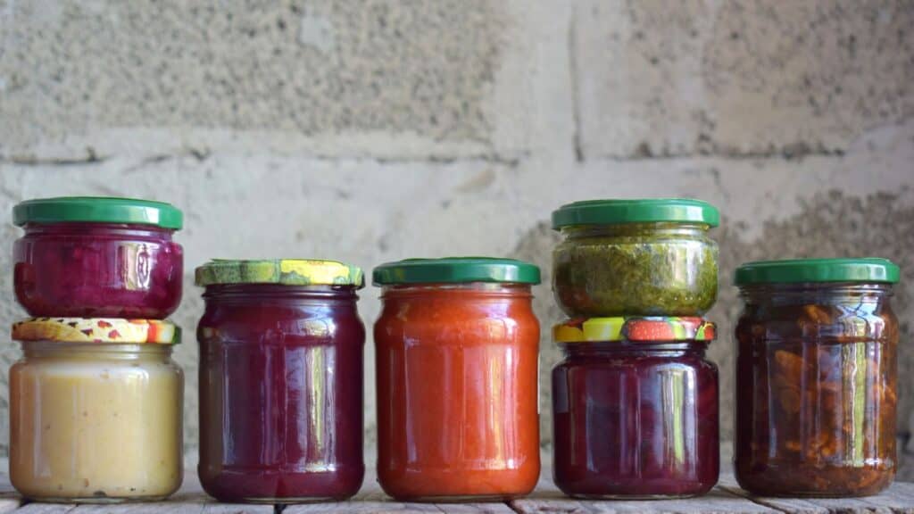 Six glass jars filled with various colorful fall harvest foods, arranged on a wooden surface against a cinder block wall.