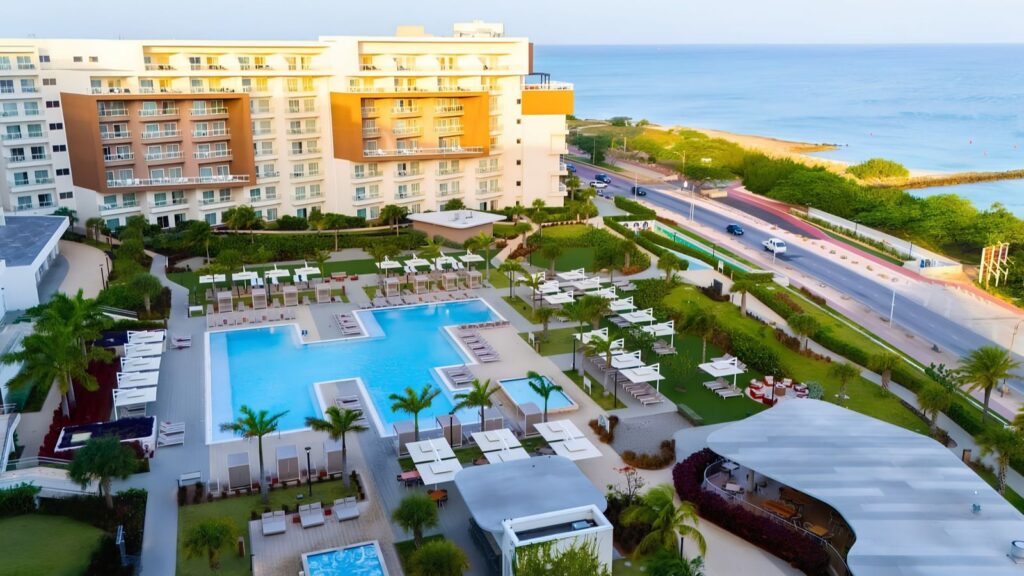 Aerial view of a beach resort hotel with a large swimming pool, lounge chairs, and an adjacent road along the coastline where Aruba draws record crowds.