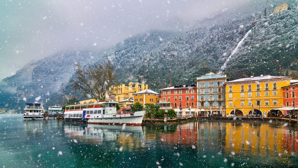 Boats docked at a colorful town waterfront during a snowfall, with snow-covered mountains in the background.