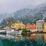 Boats docked at a colorful town waterfront during a snowfall, with snow-covered mountains in the background.