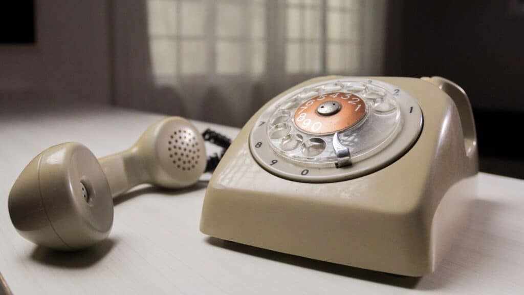 A beige rotary telephone, a landline phone model often unfamiliar to younger generations, rests with its handset off the base on a white surface.