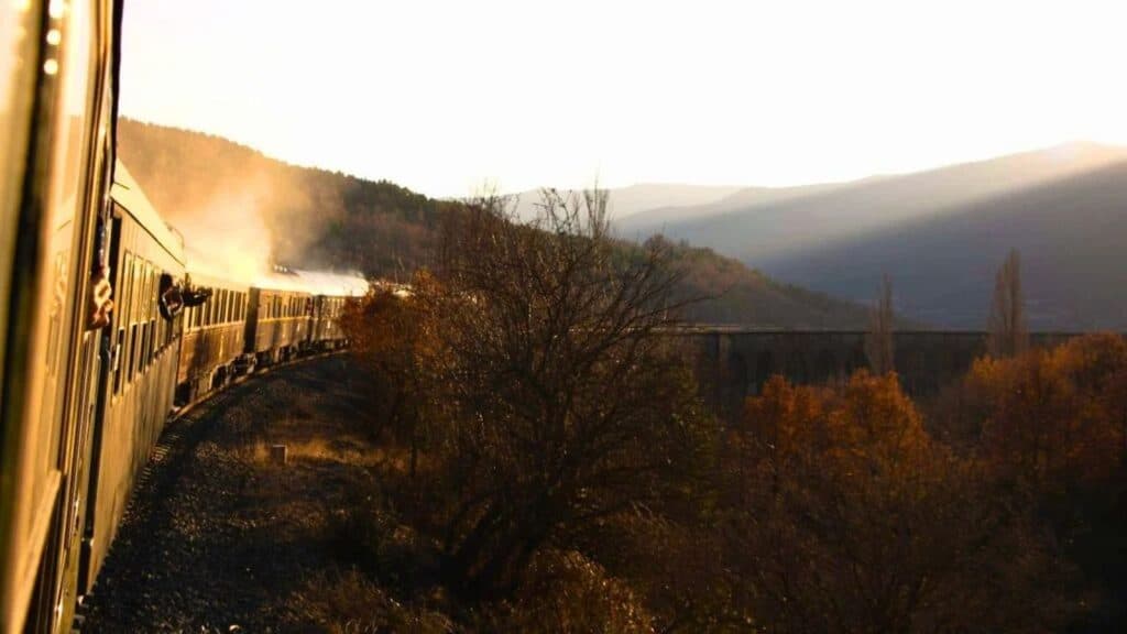 A passenger train travels along a curved track through a hilly, forested landscape filled with fall foliage at sunset.
