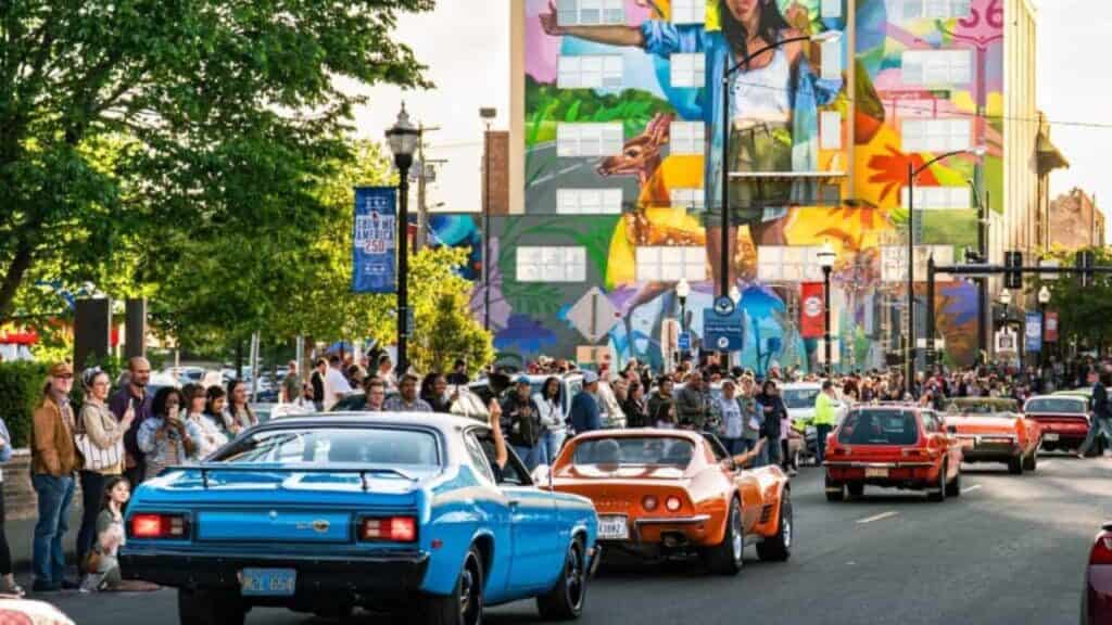 A line of classic muscle cars cruises down a city street past spectators and a mural, drawing crowds for Route 66 centennial attractions.