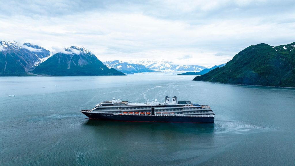 A Holland America Line cruise ship sailing through a glacial bay surrounded by mountains.