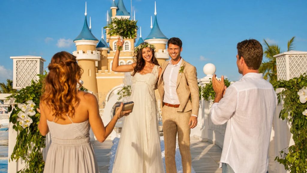 A bride and groom pose for photos in front of a castle-themed building as wedding guests capture the moment at a destination offering Caribbean wedding packages.
