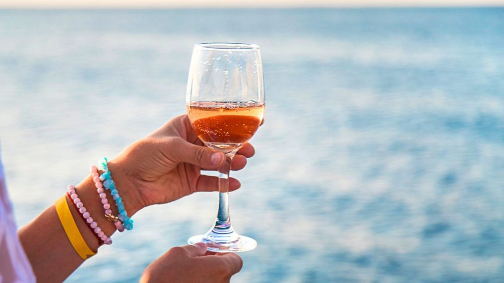 A hand holding a glass of ros&eacute; wine against a backdrop of the sea.