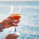 A hand holding a glass of rosé wine against a backdrop of the sea.