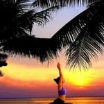 A person performs a yoga pose with arms raised overhead on a beach at sunset, framed by palm trees, evoking the peaceful atmosphere of an astrology wellness retreat.
