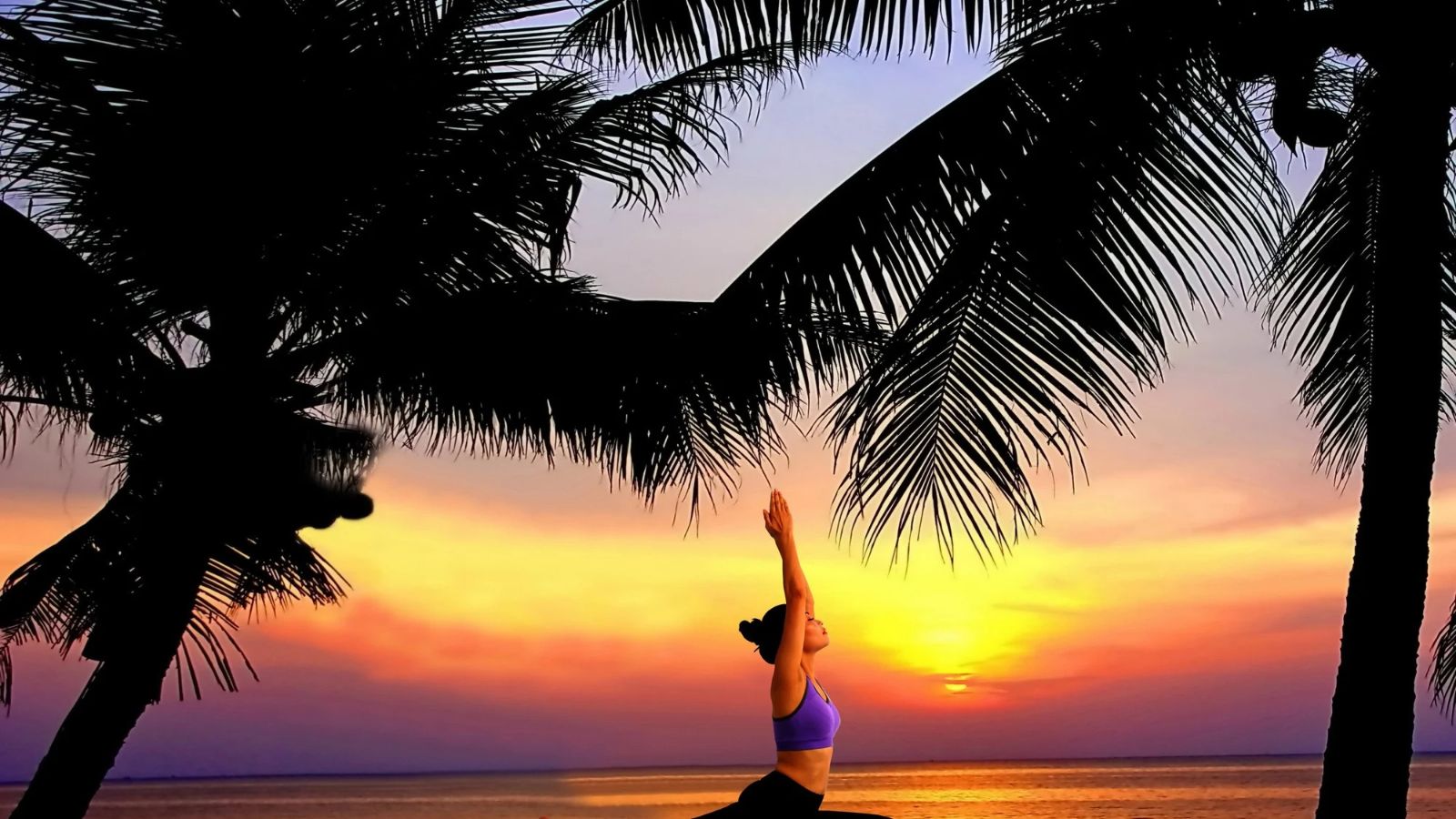 A person performs a yoga pose with arms raised overhead on a beach at sunset, framed by palm trees, evoking the peaceful atmosphere of an astrology wellness retreat.