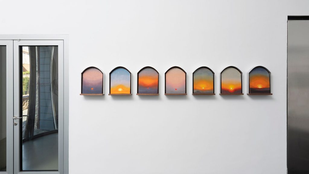Seven arched paintings of savory gelato sunset landscapes displayed in a row on a white wall.