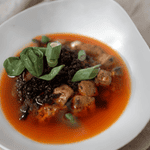 A bowl of reddish broth with chunks of meat, dark textured topping, and sliced green onions, served with a dinner roll and a spoon.