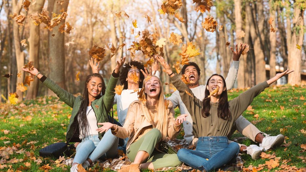 Four young adults enjoying fall activities for teens, sitting on a blanket in a park while laughing and throwing autumn leaves into the air.