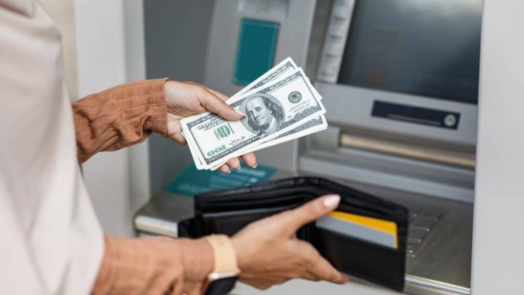 A person holds a stack of US hundred-dollar bills over an open wallet in front of an ATM, reminiscent of airport currency exchange traps.