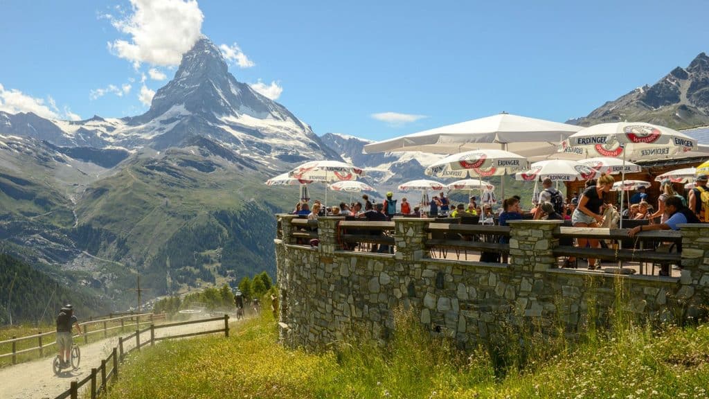 People seated at an outdoor restaurant terrace with the Matterhorn in the background, as a person on a mountain scooter rides along a nearby dirt path.
