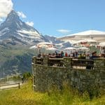 People seated at an outdoor restaurant terrace with the Matterhorn in the background, as a person on a mountain scooter rides along a nearby dirt path.
