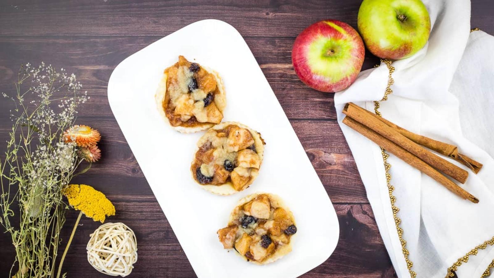 Three small apple and raisin tarts on a rectangular plate, surrounded by two apples, cinnamon sticks, and dried flowers on a wooden table.
