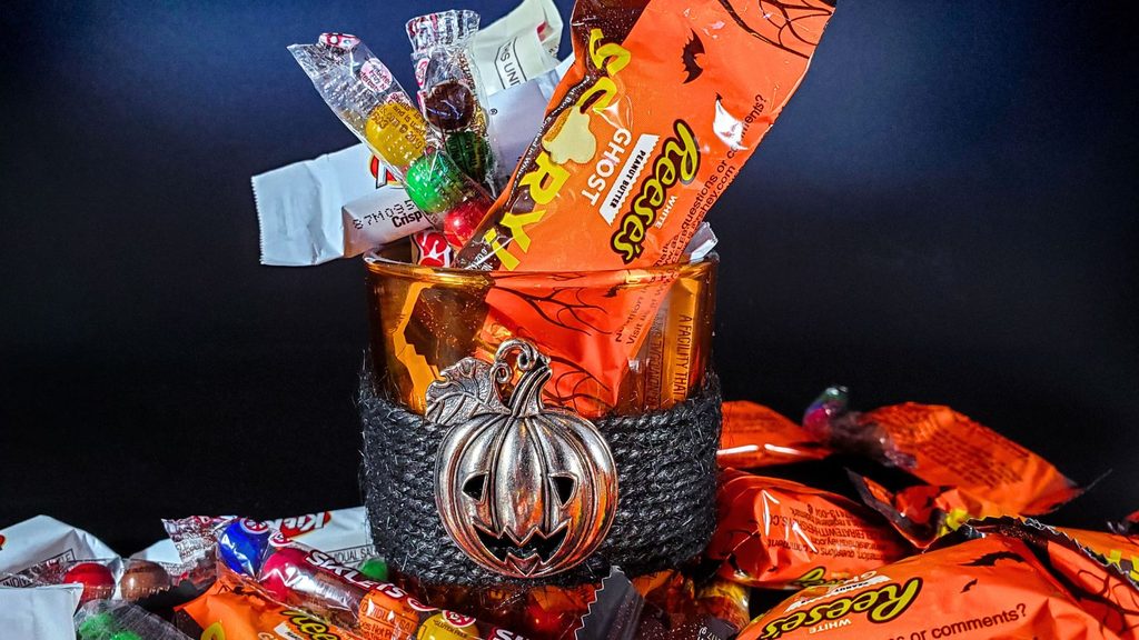 A glass votive holder decorated with a metal pumpkin charm, overflowing with assorted National Chocolate Day candy.