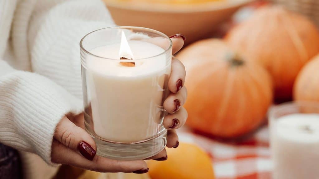 A person holds a lit fall candle in a glass jar, with pumpkins in the blurred background.