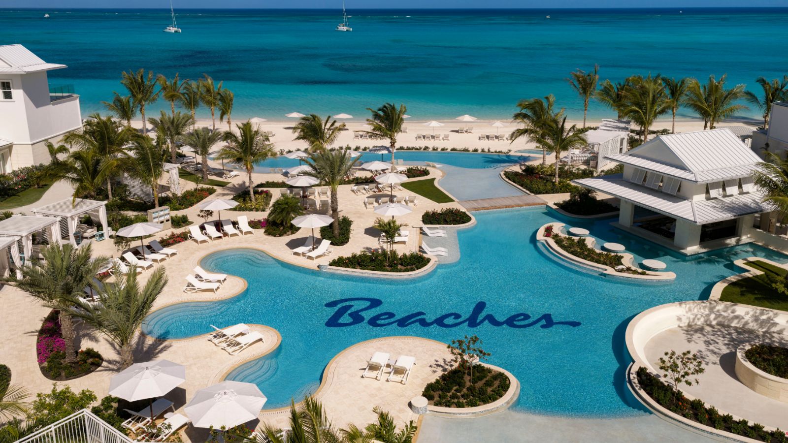 Aerial view of a Royal Caribbean Group luxury resort pool complex with the &ldquo;Beaches&rdquo; logo on the floor, surrounded by palm trees and beach chairs, overlooking the ocean.