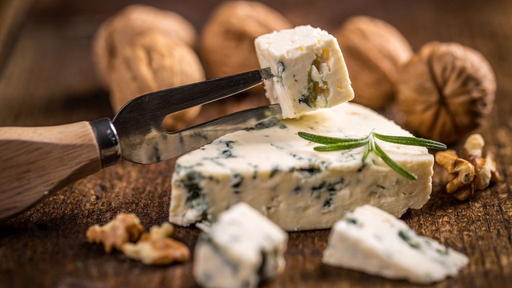 A cheese fork holds a cube of blue cheese for National Moldy Cheese Day above a wedge garnished with a rosemary sprig, surrounded by whole walnuts on a wooden surface.