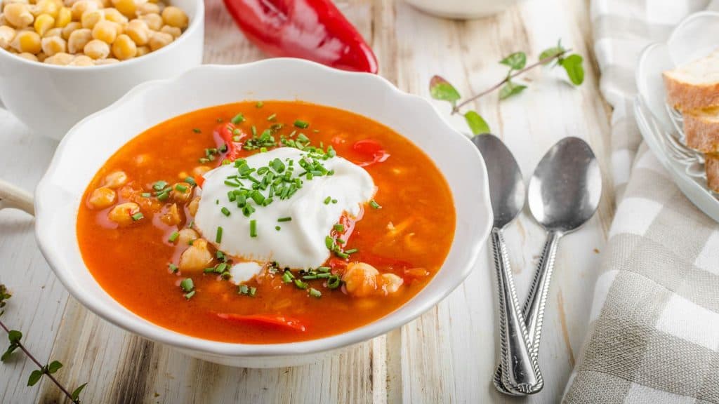 A bowl of chickpea soup with red pepper, topped with a dollop of sour cream and chives, served with a side of chickpeas and bread.