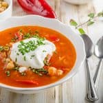A bowl of chickpea soup with red pepper, topped with a dollop of sour cream and chives, served with a side of chickpeas and bread.