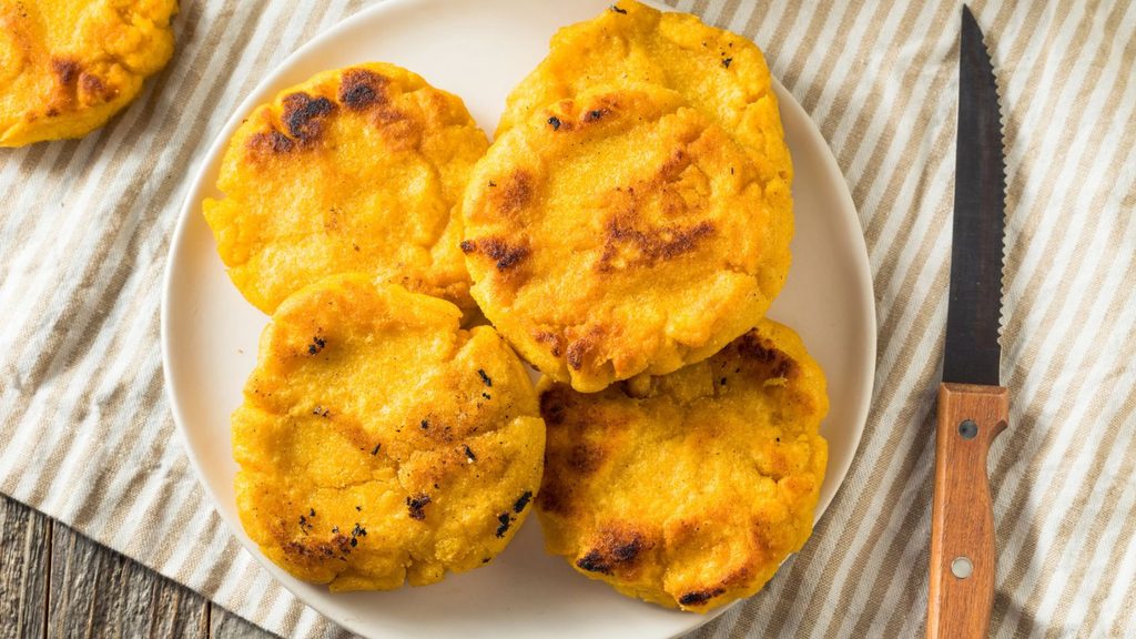 Four golden-yellow corn arepas on a white plate next to a steak knife.