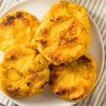 Four golden-yellow corn arepas on a white plate next to a steak knife.