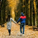A man and a woman holding hands walk away from the camera down an autumn park path lined with trees and fallen leaves.