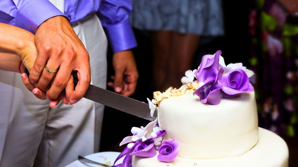 A couple&rsquo;s hands holding a knife to cut a white wedding cake decorated with purple sugar roses.