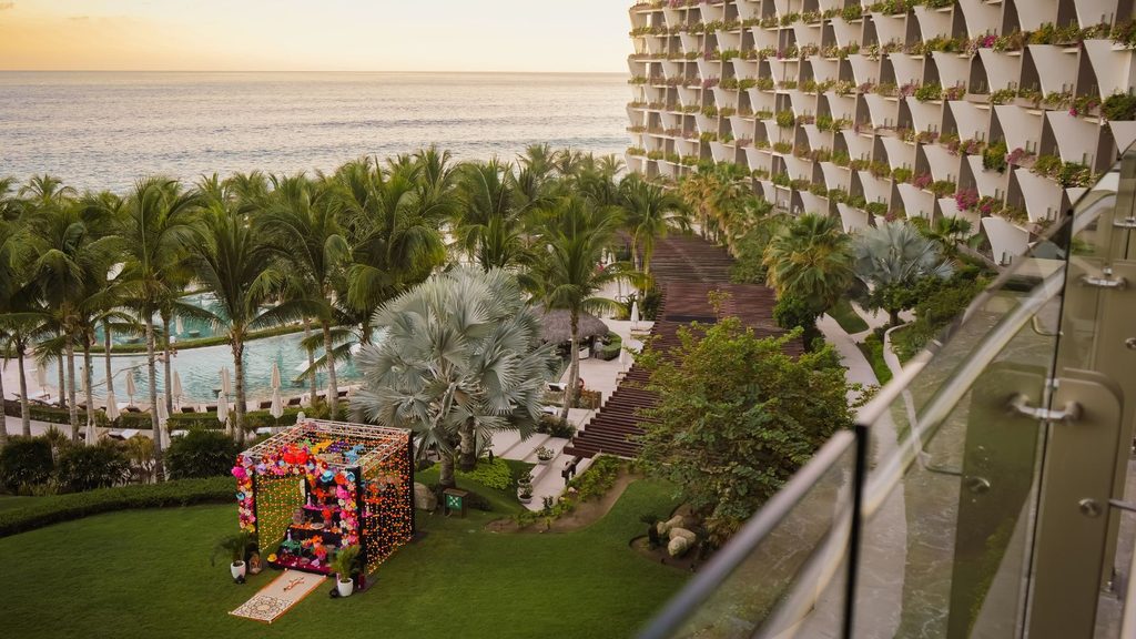 An elevated view of a resort with a large tiered hotel building, palm trees, a swimming pool, and a decorated altar set up on a lawn overlooking the ocean.
