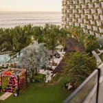 An elevated view of a resort with a large tiered hotel building, palm trees, a swimming pool, and a decorated altar set up on a lawn overlooking the ocean.