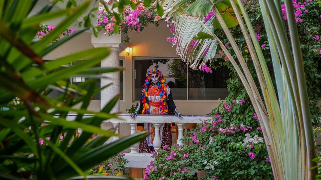 A person wearing Catrina makeup and a colorful traditional outfit stands on a balcony framed by tropical foliage and bougainvillea flowers.