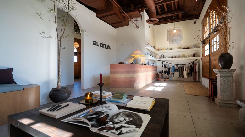 Interior of a boutique store featuring an arched entryway, a layered clay-colored service counter, and a coffee table with open books in the foreground.