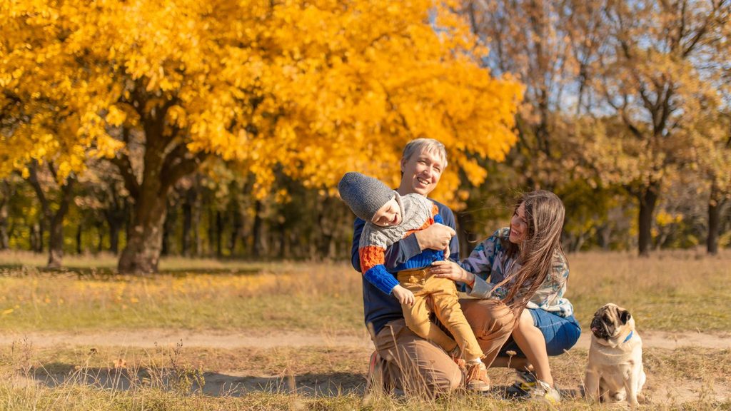 A smiling couple plays with a young child among golden autumn trees in a park, a perfect thing to do in October as a pug sits nearby.