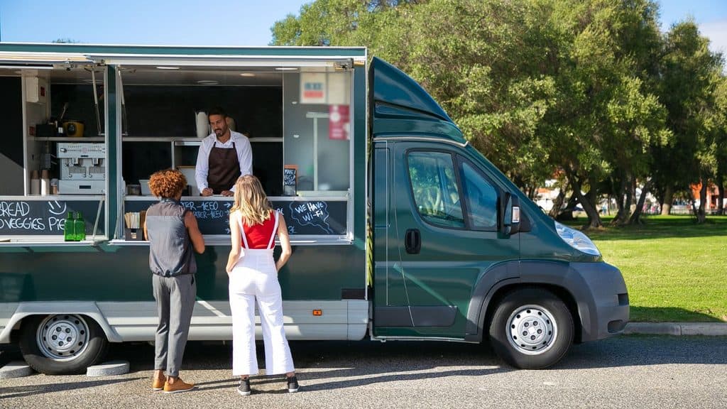A food truck vendor waits as two customers consider tipping after their service at an outdoor parking area.