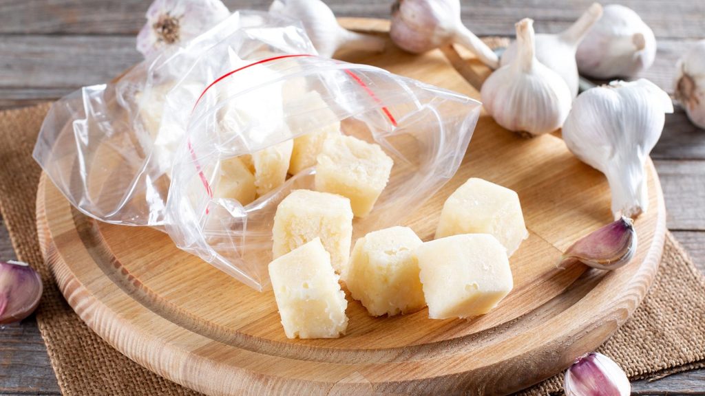 Frozen garlic cubes in a plastic bag on a wooden cutting board with whole garlic bulbs.