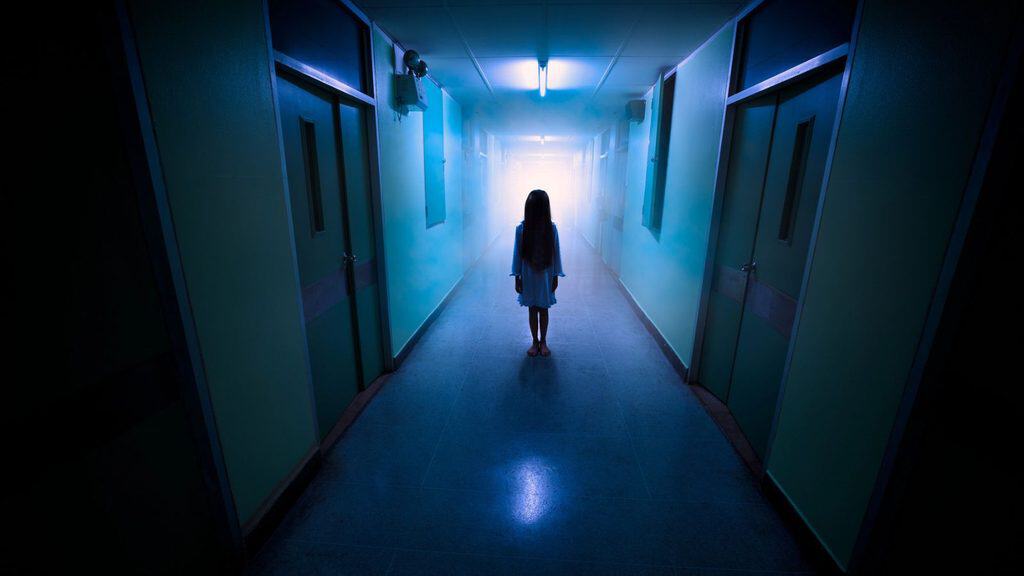A girl in a white dress stands at the end of a dimly lit, blue-toned hospital corridor with bright light from a doorway, perfect for halloween travel imagery.