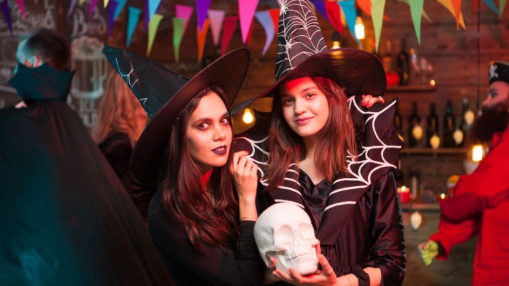 Two young women in witch costumes at an adult Halloween party, with one holding a plastic skull.
