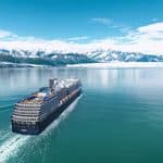 A cruise ship sailing on a calm bay in front of a glacier and snow-capped mountains.