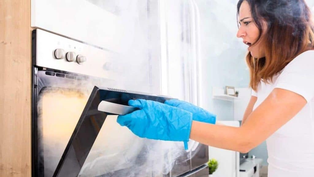 A woman wearing blue oven mitts opens an oven door, releasing a cloud of smoke.