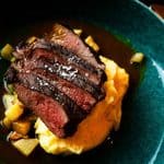 Sliced seared steak topped with sea salt, served over mashed potatoes and sautéed vegetables in a dark sauce.