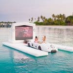 Two people sit in a white sports car positioned on a floating platform with a large inflatable movie screen in the ocean.