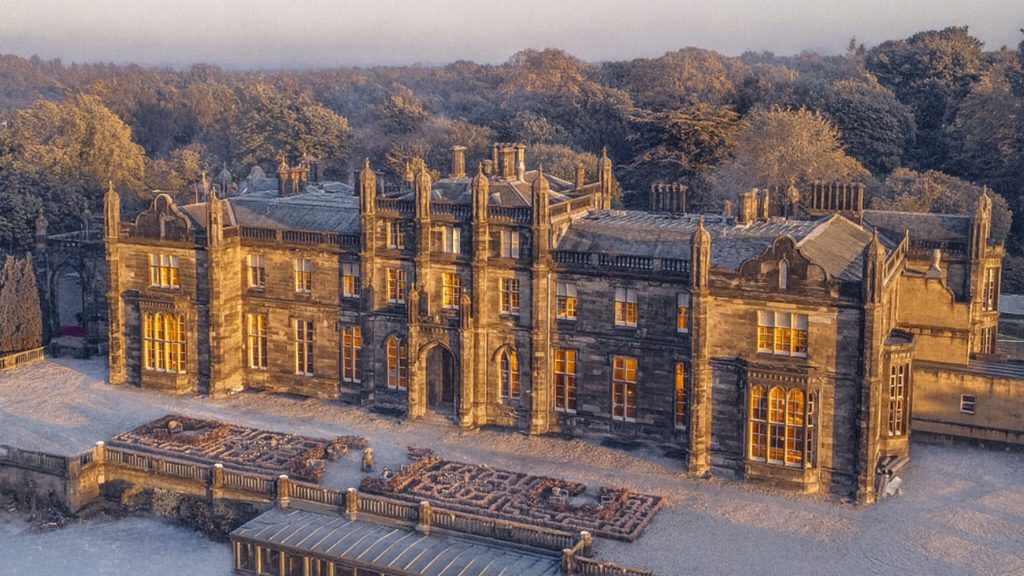 Aerial view of a large stone mansion with lit windows, situated next to a garden maze and surrounded by trees.