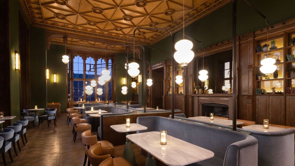 A restaurant interior with dark wood paneling, paneled ceilings, and rows of blue booths with marble-topped tables illuminated by modern glass pendant lights.